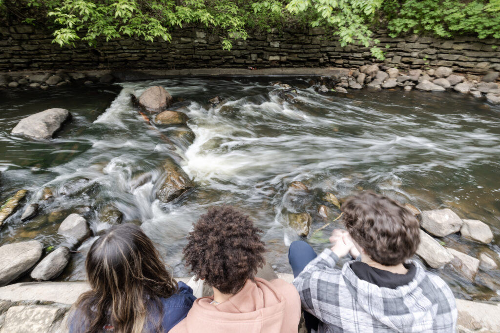 Three teens, their backs to the camera, sit on rocks along a rushing river.