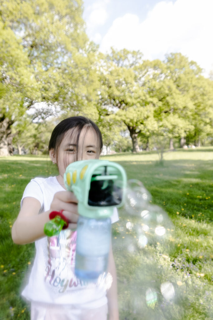 A young girl with black bangs in her face shoots a bubble gun toward the camera.