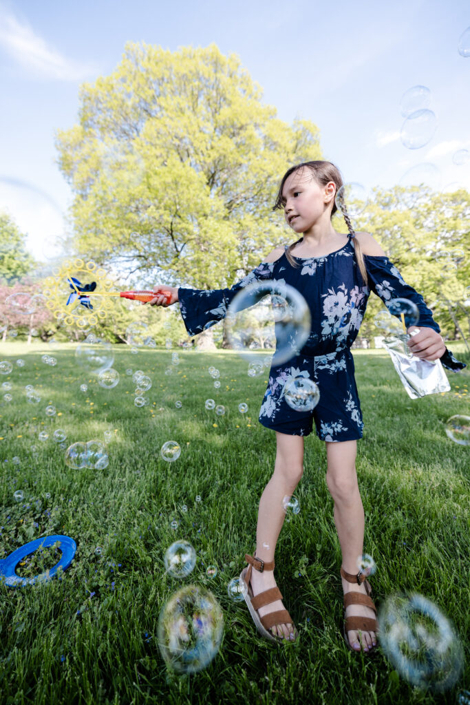 A young girl with two braids in a blue flowered romper and brown sandals shoots bubbles toward the camera.