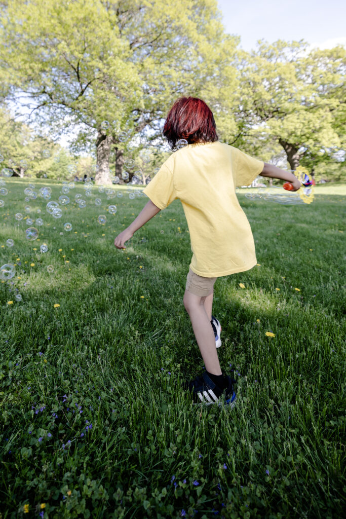 A child with short red hair in a yellow shirt runs through the grass, turned away from the camera with a bubble gun.