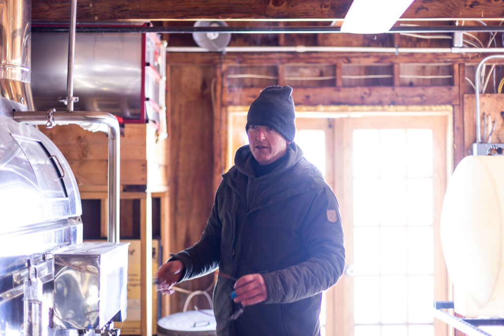 A man in a dark green jacket stands inside a maple sugar house where syrup is made from tree sap.