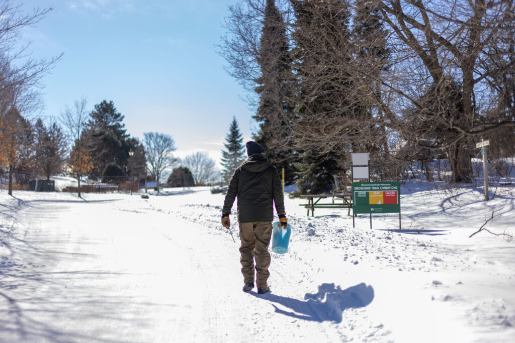 A man in a dark green jacket and tan pants walks down a snowy path away from the camera on a sunny winter day.