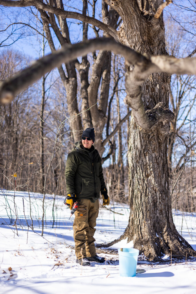 A man in a dark green jacket and tan pants stands in a forest in winter.