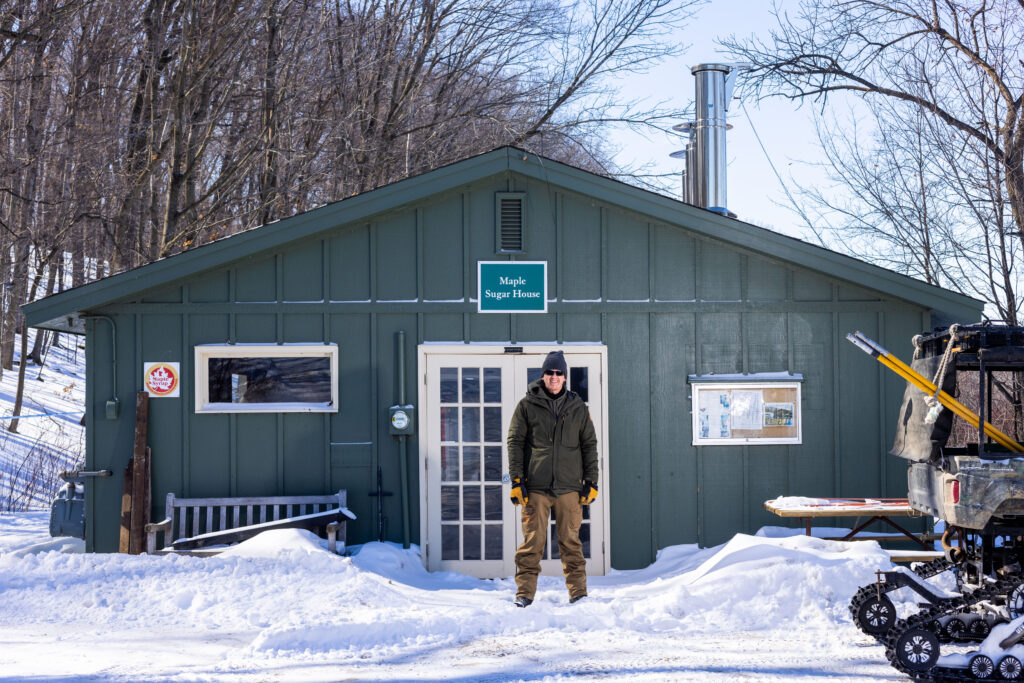 A man in a dark green jacket stands outside a maple sugar house where syrup is made from tree sap.