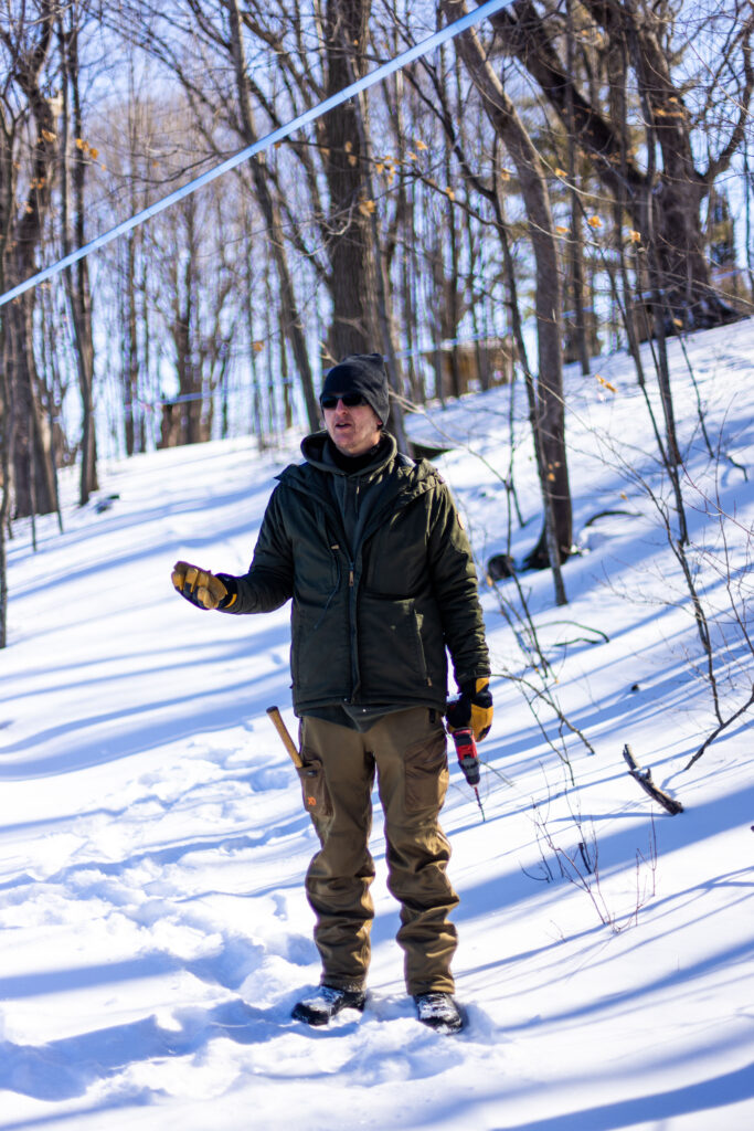 A man in a dark green jacket and tan pants stands in a forest in winter, explaining to his audience.