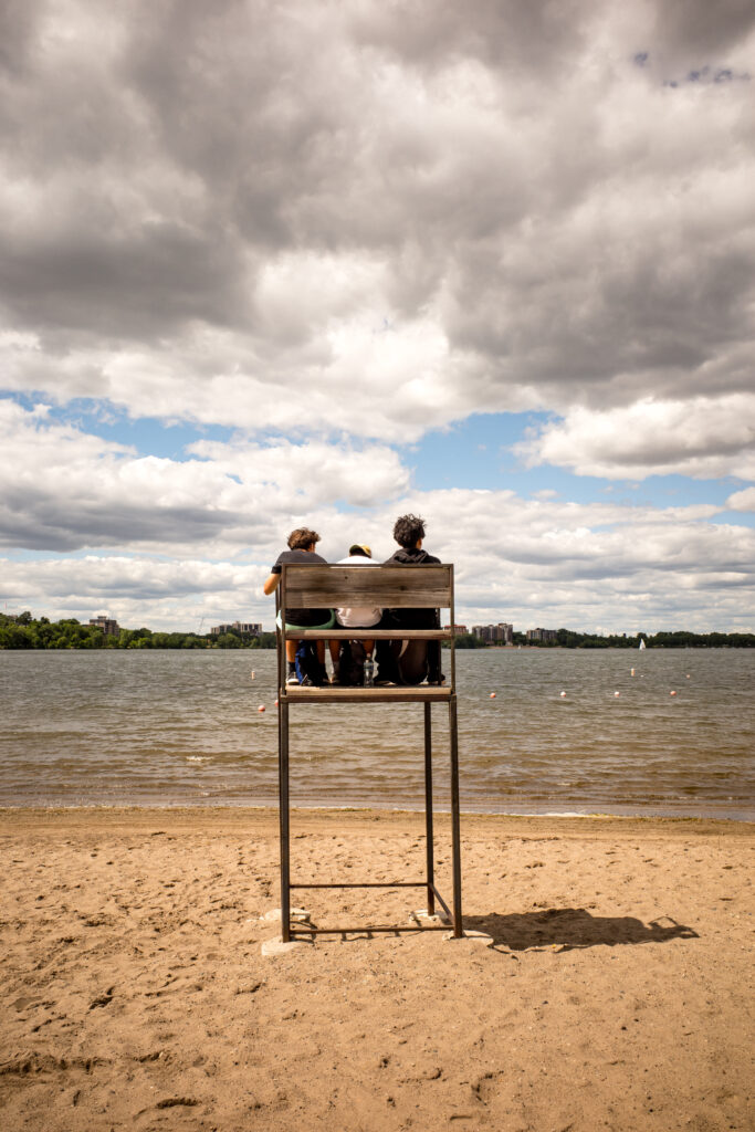 Three people sit on a raised lifeguard chair while looking out over a lake.
