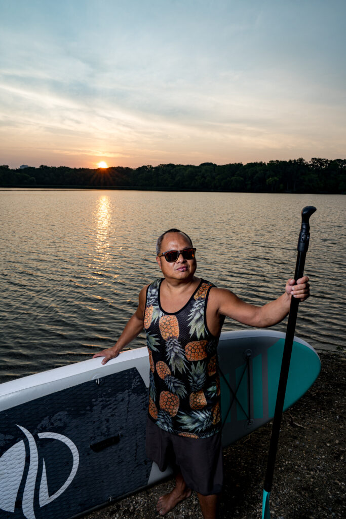 A man with a gray ponytail and wearing a tank top covered in pineapples stands facing the camera the camera holding a paddleboard and an oar.