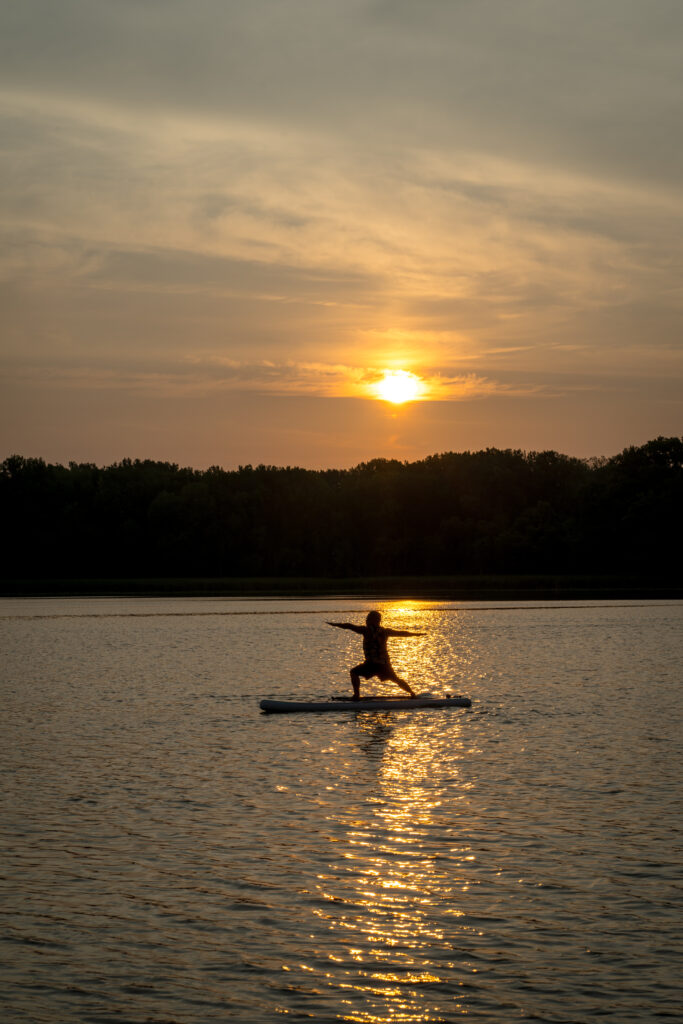 The silhouette of someone doing warrior pose on a board in the middle of a lake while the sun rises.
