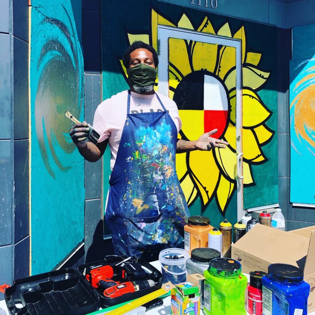 A person in a paint-splattered apron, white shirt, and facemask standing next to paint in front of a mural of a yellow flower.