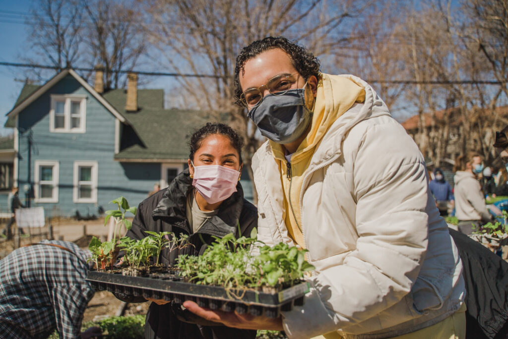 Two smiling people, one in a puffy white coat and the other in a black one, hold up a box of seedlings happily.