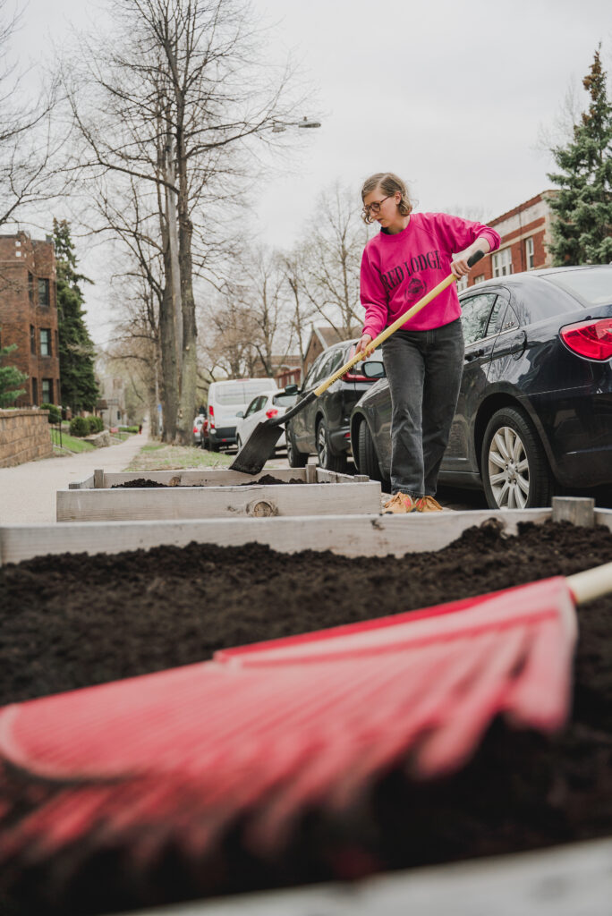 A person in a bright pink sweatshirt uses a garden tool to prep dirt in a raised-bed garden.
