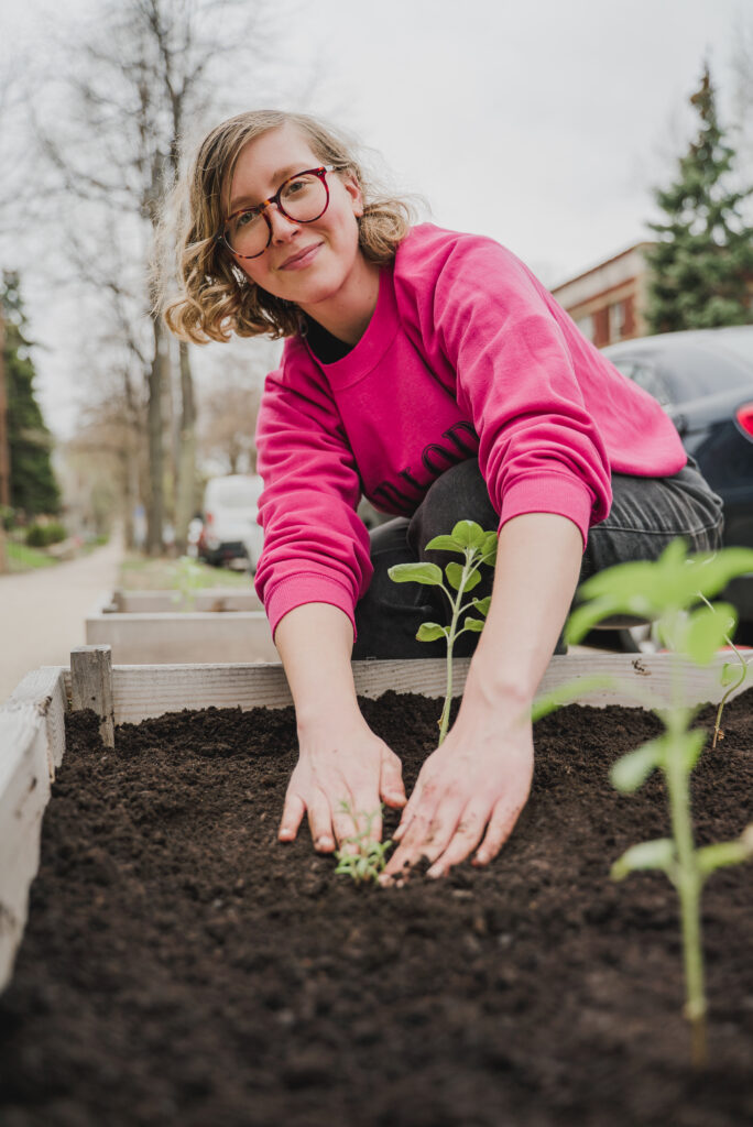 A woman with glasses in a bright pink sweatshirt plants new seedlings in rich dirt in a raised-bed garden plot.