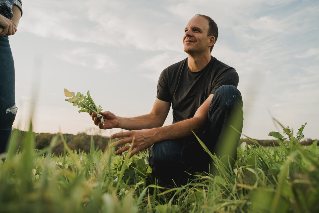 A man in a black t-shirt kneels in a field and holds a young crop in his hand.