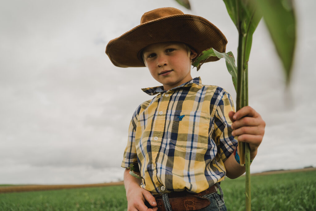 A young boy in a plaid yellow shirt and cowboy hat looks at the camera with a sprout in his hand.