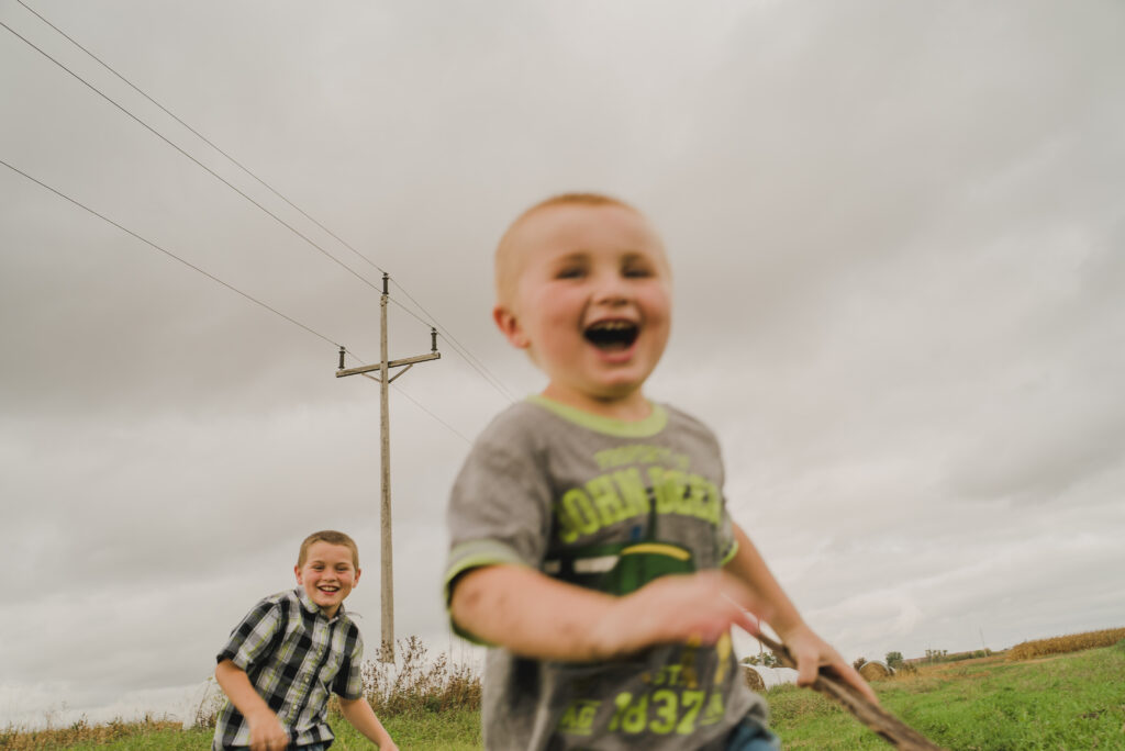 Two young boys run in a field, the younger one in a gray t-shirt close to the camera laughing.
