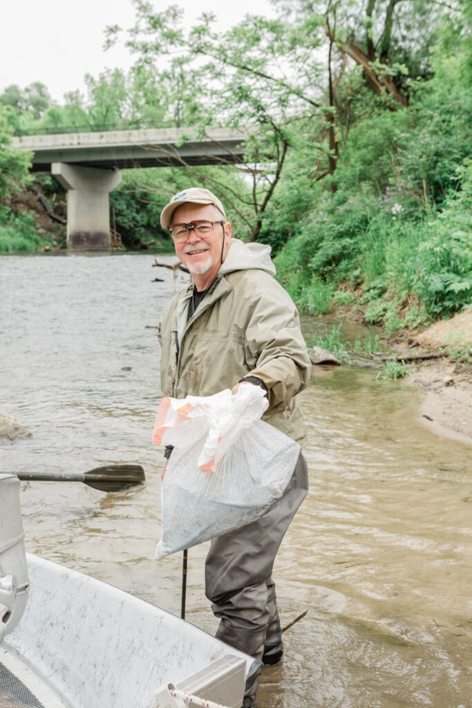An older man in glasses, a hat, and tan fishing gear holds up a plastic garbage bag filled with trash with a smile while he stands in the river near a bank.
