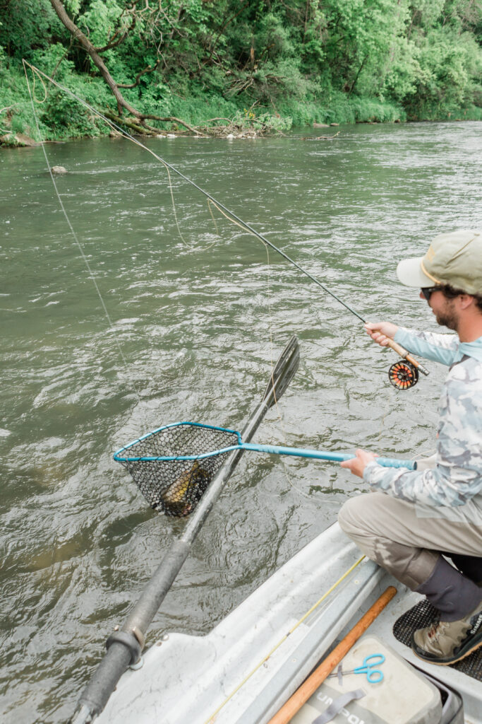A man squats on his boat, fishing rod in one hand and a net with a fish in it in the other.