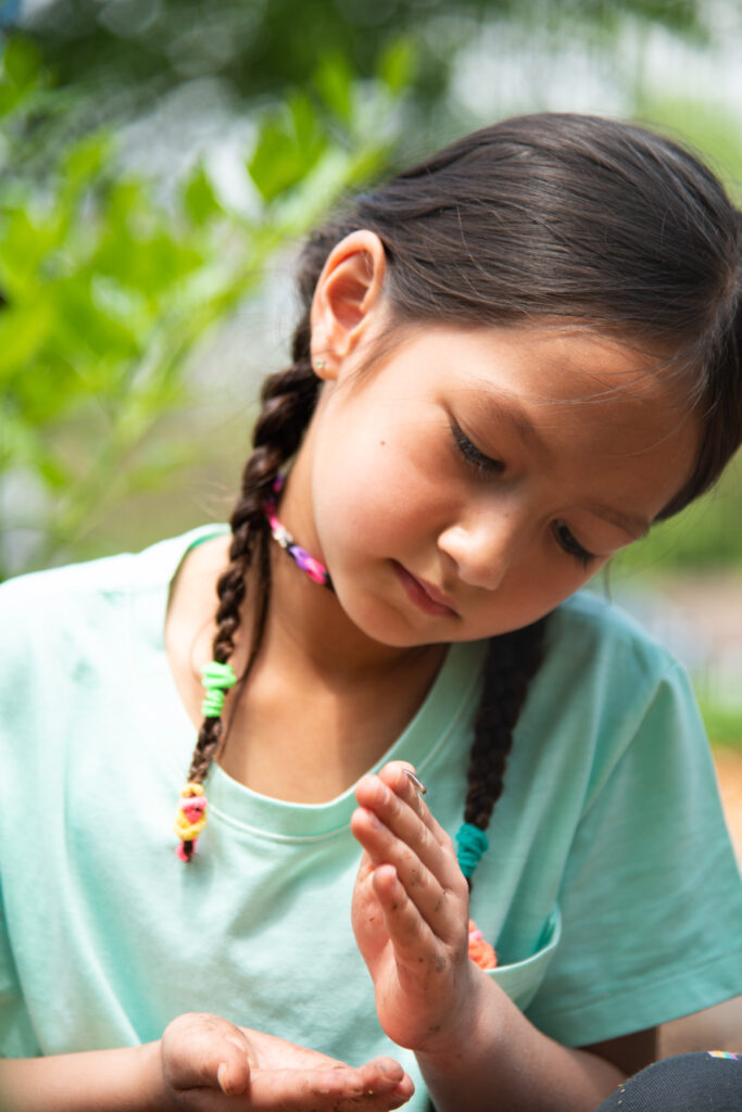 A closeup of a young girl with two braids in a blue shirt holding her hands up and inspecting a small worm that is crawling on them.