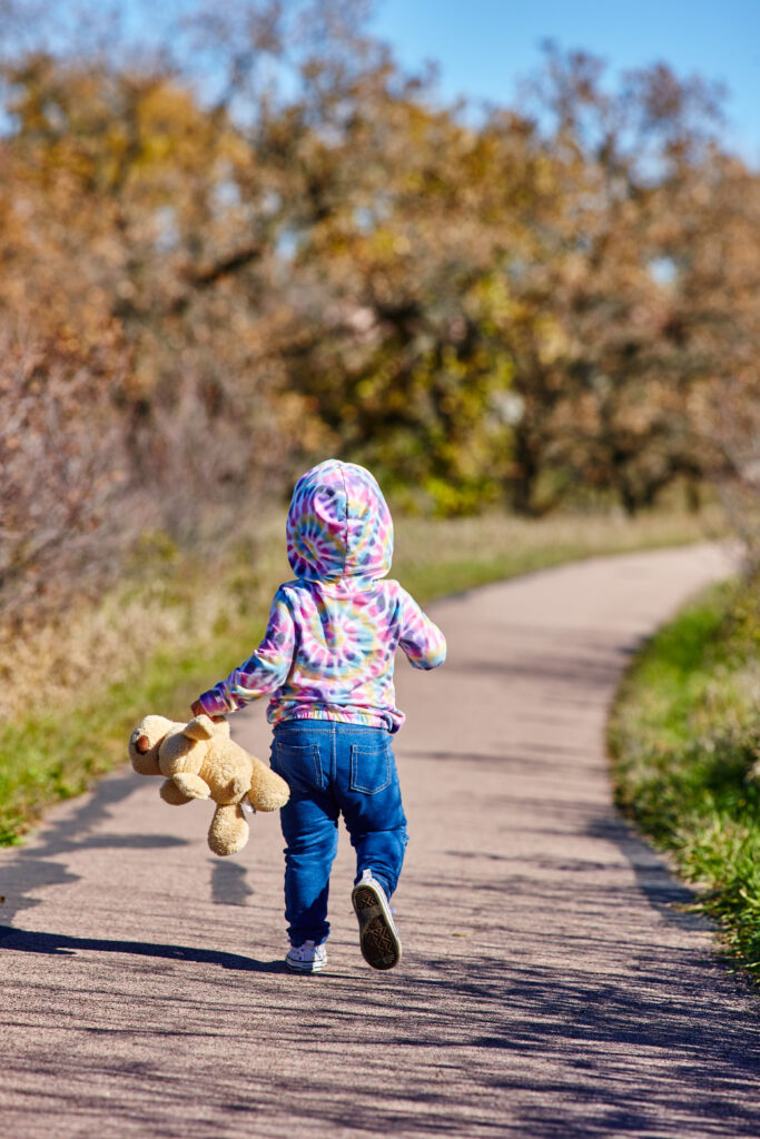 A child in blue pants and a pink and blue hoodie holding onto a stuffed teddy bear runs down a path away from the camera.