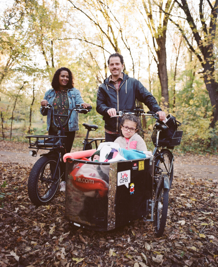 A family of three with a young girl in a bike wagon next to her parents who stand near their bikes.