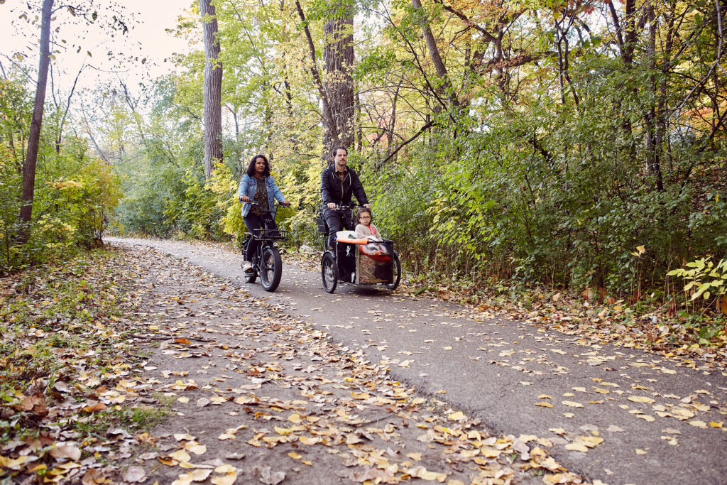 A family of three, a young girl in a bike trailer and two adults on their bikes, bike toward the camera down a bike path.