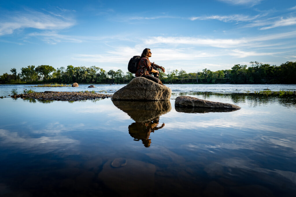 A man with shoulder-length hair and a backpack sits on a rock in the middle of a lake and looks out over the water.