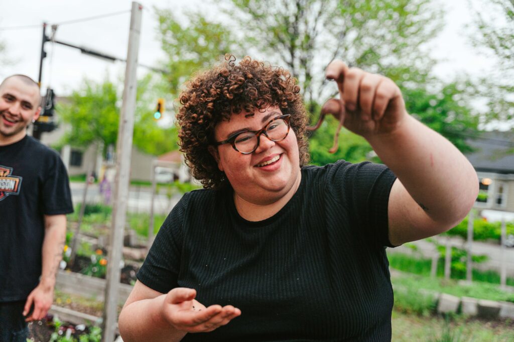 A person with short curly hair, black glasses, and a black t-shirt holds up a worm in their fingers.