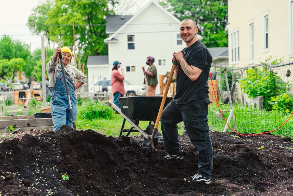 Two people, one in a yellow hat and blue overalls and one in all black, pose holding shovels by a large pile of dark dirt.