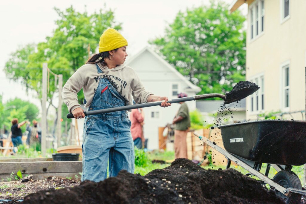A person in a yellow hat and blue overalls tosses a shovelful of dirt.