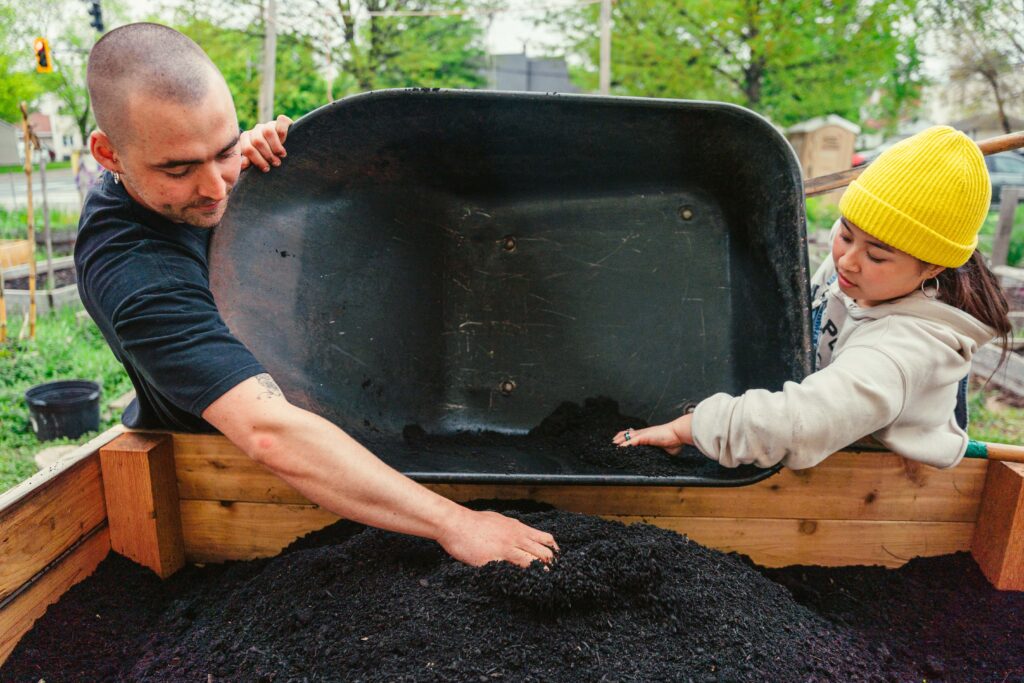 Two people, one in a yellow hat and blue overalls and one in all black, dump a black wheelbarrow of dirt into a wooden box.