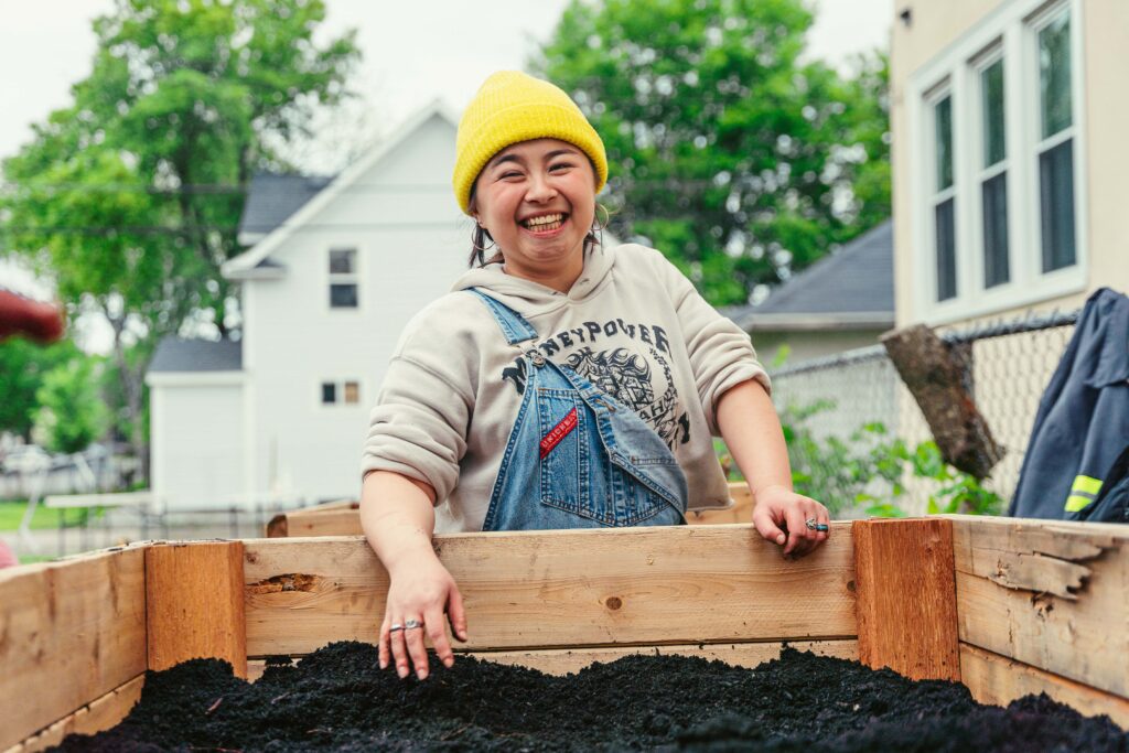 A person in a yellow hat and blue overalls smiles with their hand in a wooden box filled with dirt.