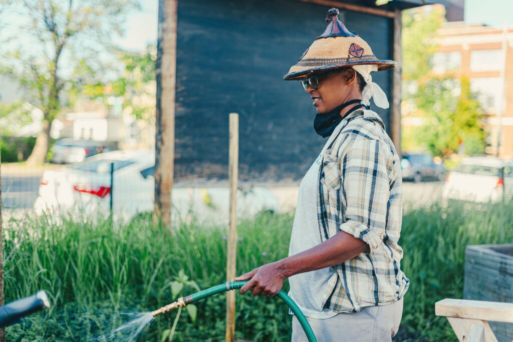 A person in a pointed hat and white plaid shirt holds a hose and waters a garden ;ot.