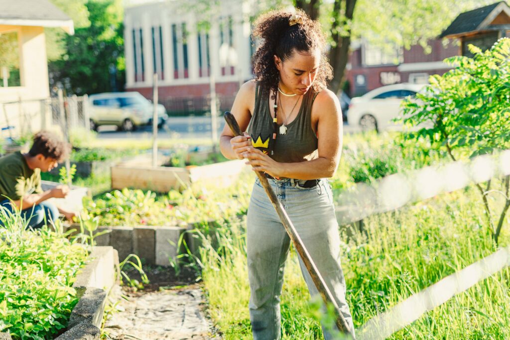 A person with dark curly hair tied up behind their head holds a garden tool next to a garden plot.