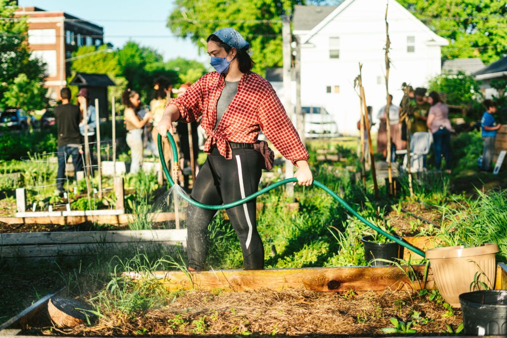 A person with a blue bandana, a red shirt, and black pants holds a hose and waters a garden plot.