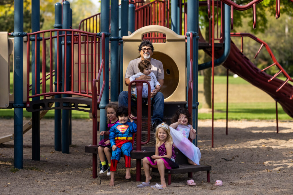 A man with dark hair and a white beard sits on a playground with a young child in his lap and four children sitting on steps below him smiling.