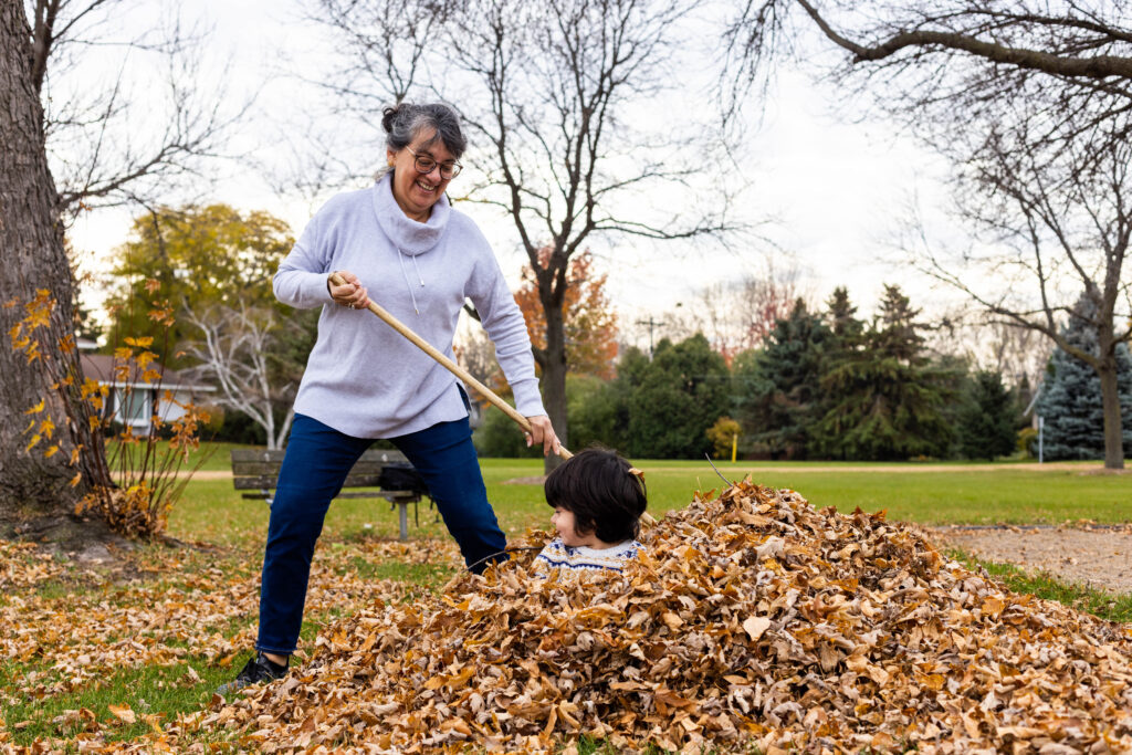A person in jeans and a gray sweater rakes brown leaves into a large pile where a young child sits among the leaves.