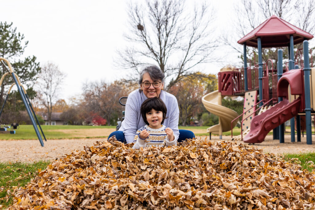 An older woman in a gray sweater holds a young boy in a pile of leaves and smiles at the camera.