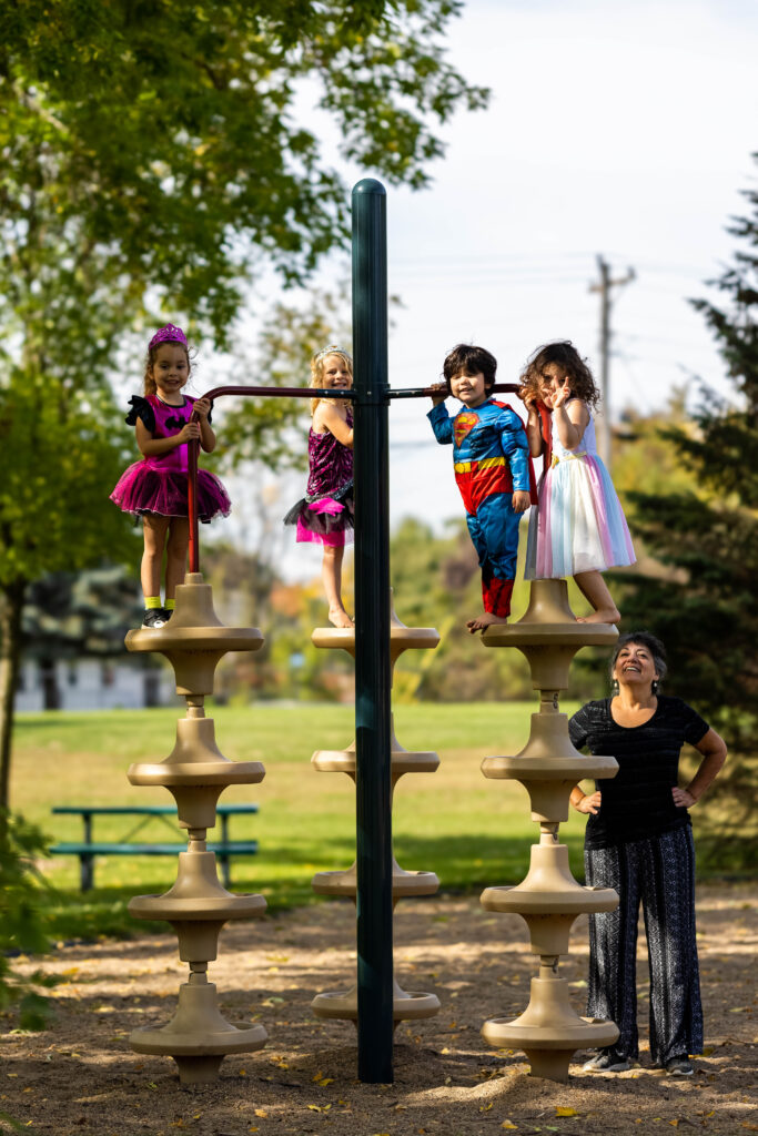 Four children, one in a Superman costume and one in a bright pink and black dance costume, stand on a jungle gym while a woman in all black watches them smiling.