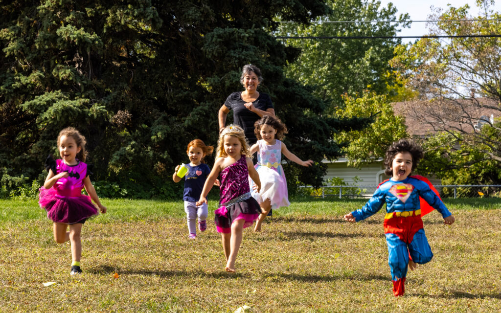 Four children, one in a Superman costume and one in a bright pink and black dance costume, run on a grassy field toward the camera, while a woman in all black watches them smiling.