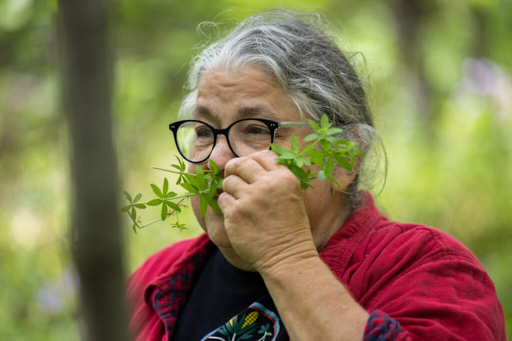 A woman in a red shirt with a black t-shirt underneath holds a plant up to her nose to smell.