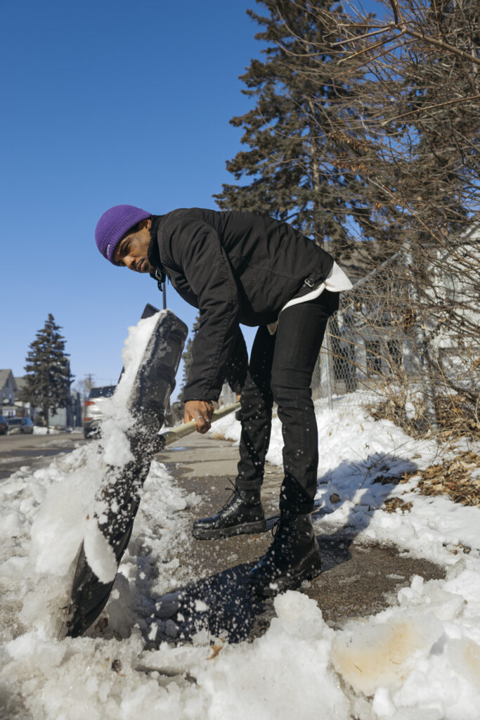 A man in all black with a purple hat shovels snow off a sidewalk on a sunny day.