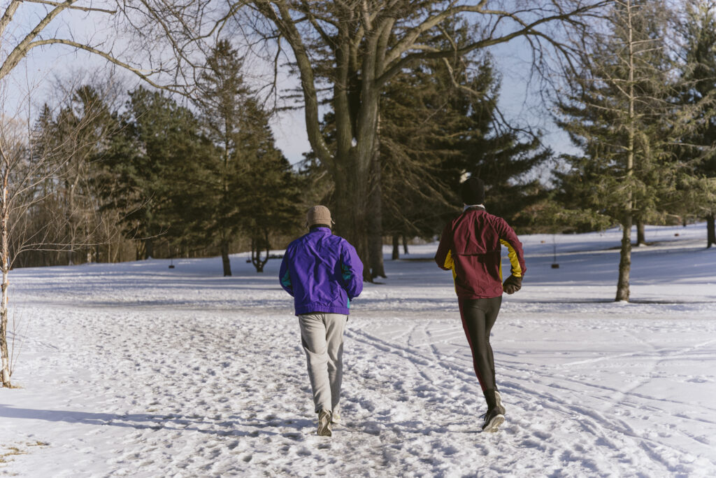 Two people, one in purple and gray and one in red and black, run through the snow away from the camera.
