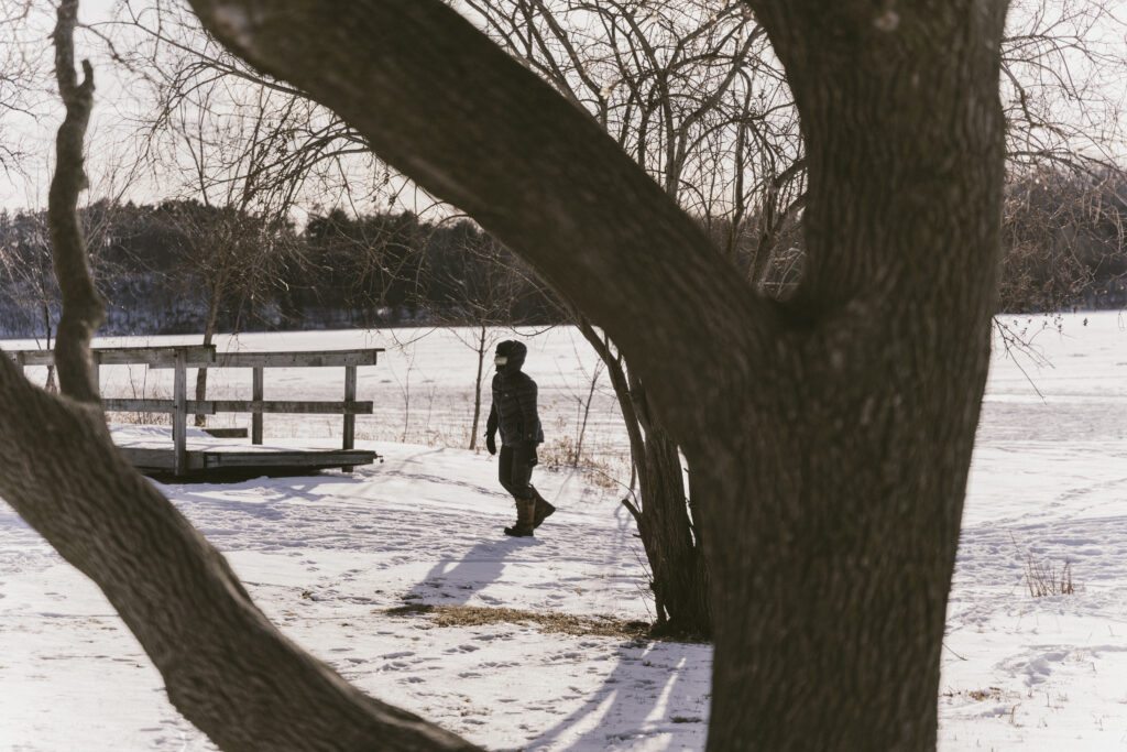 A bundled up person walking through the snow can be seen through the branches of a tree.