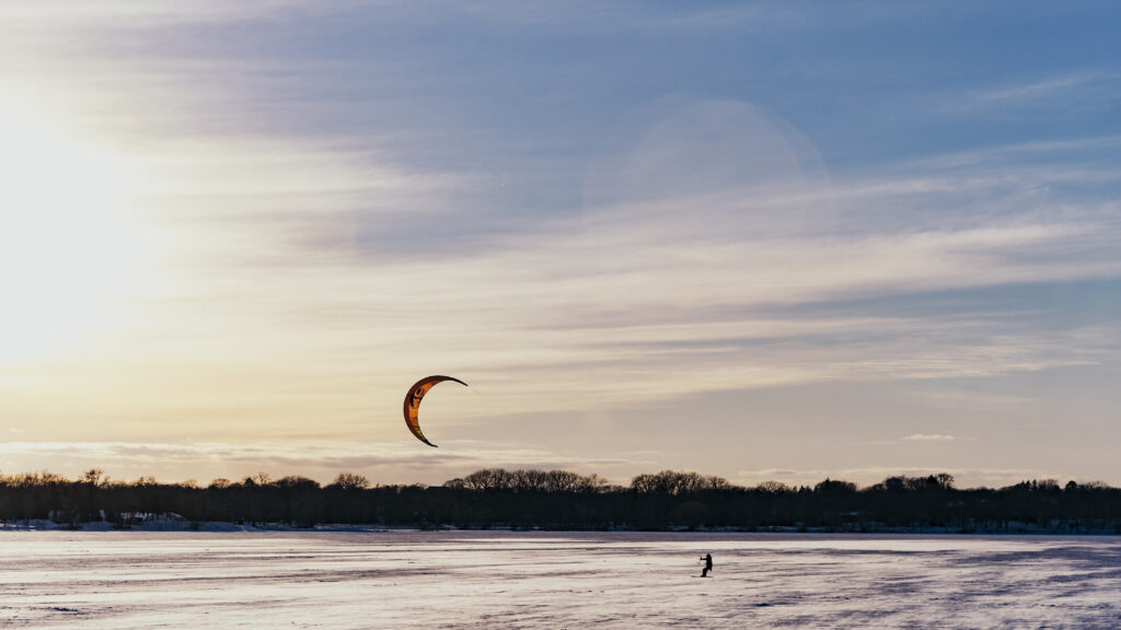 In the distance on a frozen lake, a small person can be seen being pulled by a large orange kite.