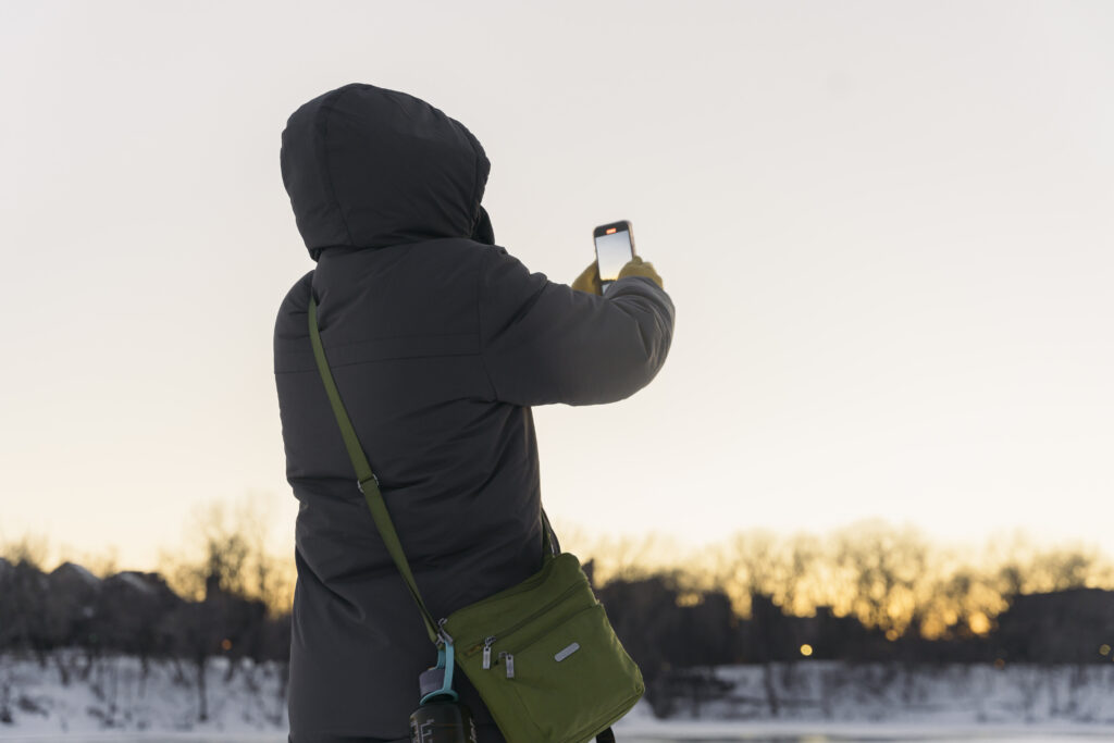 A person in a gray coat with a green bag around their shoulders holds their phone up to take a picture of a snowy park.