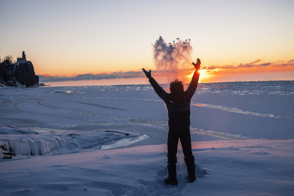 The silhouette of a person throwing snow above their head in front of a rising sun.