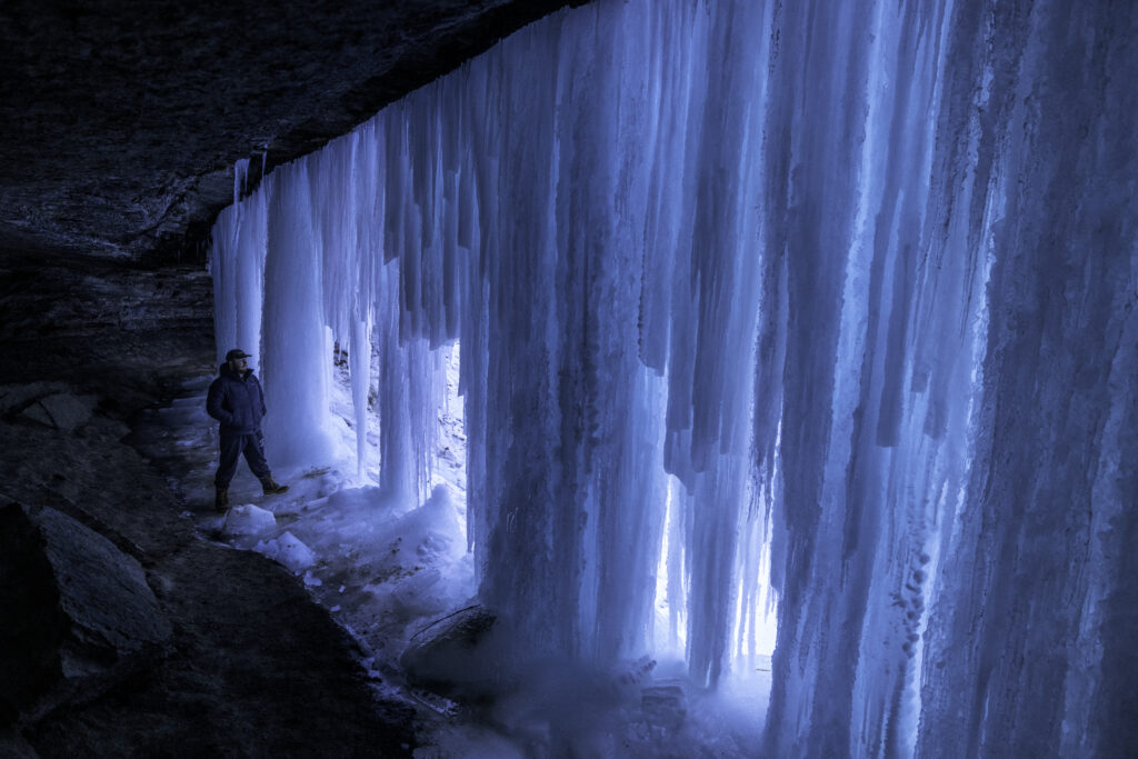 Large icicles block the entrance to a cave where a person, dwarfed by the ice, stands behind them.