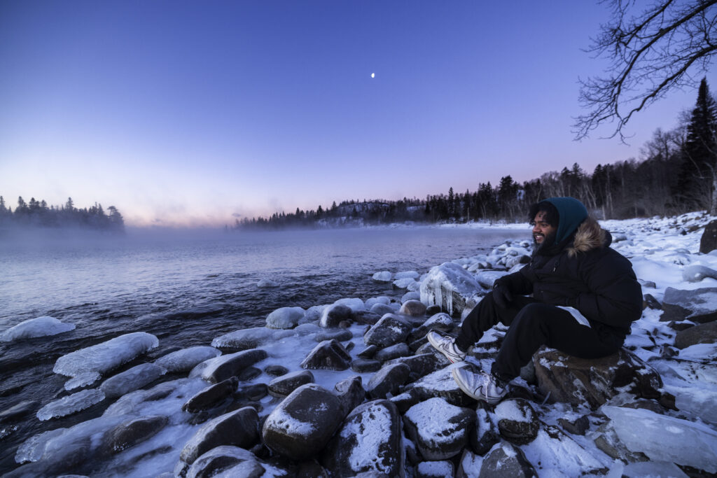 A person in a blue hoodie and black jacket sits on snowy rocks and looks out at steam rising from the lake on a winter evening.