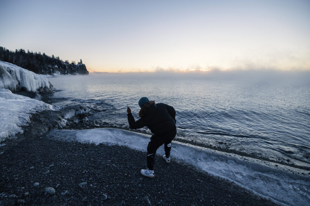 A person in dark clothing bends over as if skipping a stone across a steamy lake on a winter day.