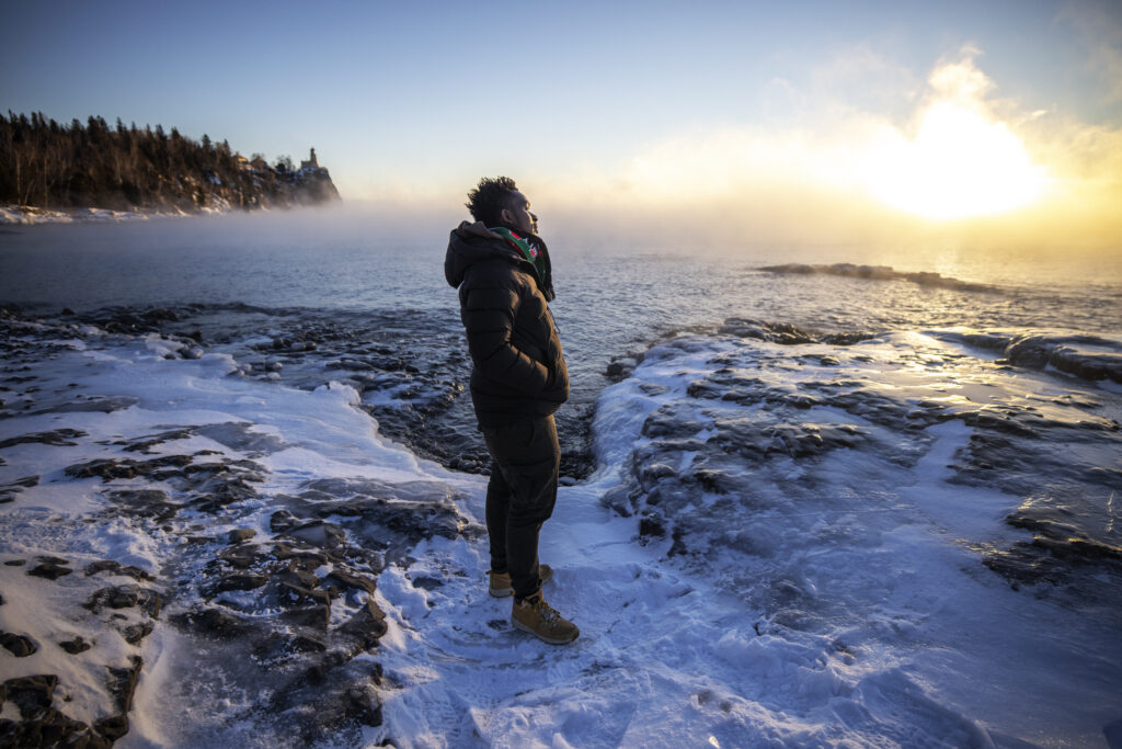 A person in dark clothing stands on a patch of snow overlooking a cold lake in pale sunlight.